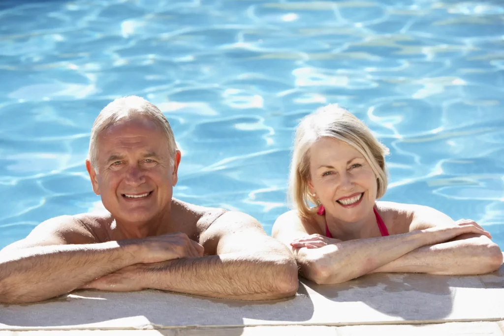 A smiling senior couple in an inground pool stand at the water's edge with their arms folded on the ledge of the pool deck. Their ability to secure themselves at the pool's edge serves the discussion of why in-pool chairs are easy for seniors to use.