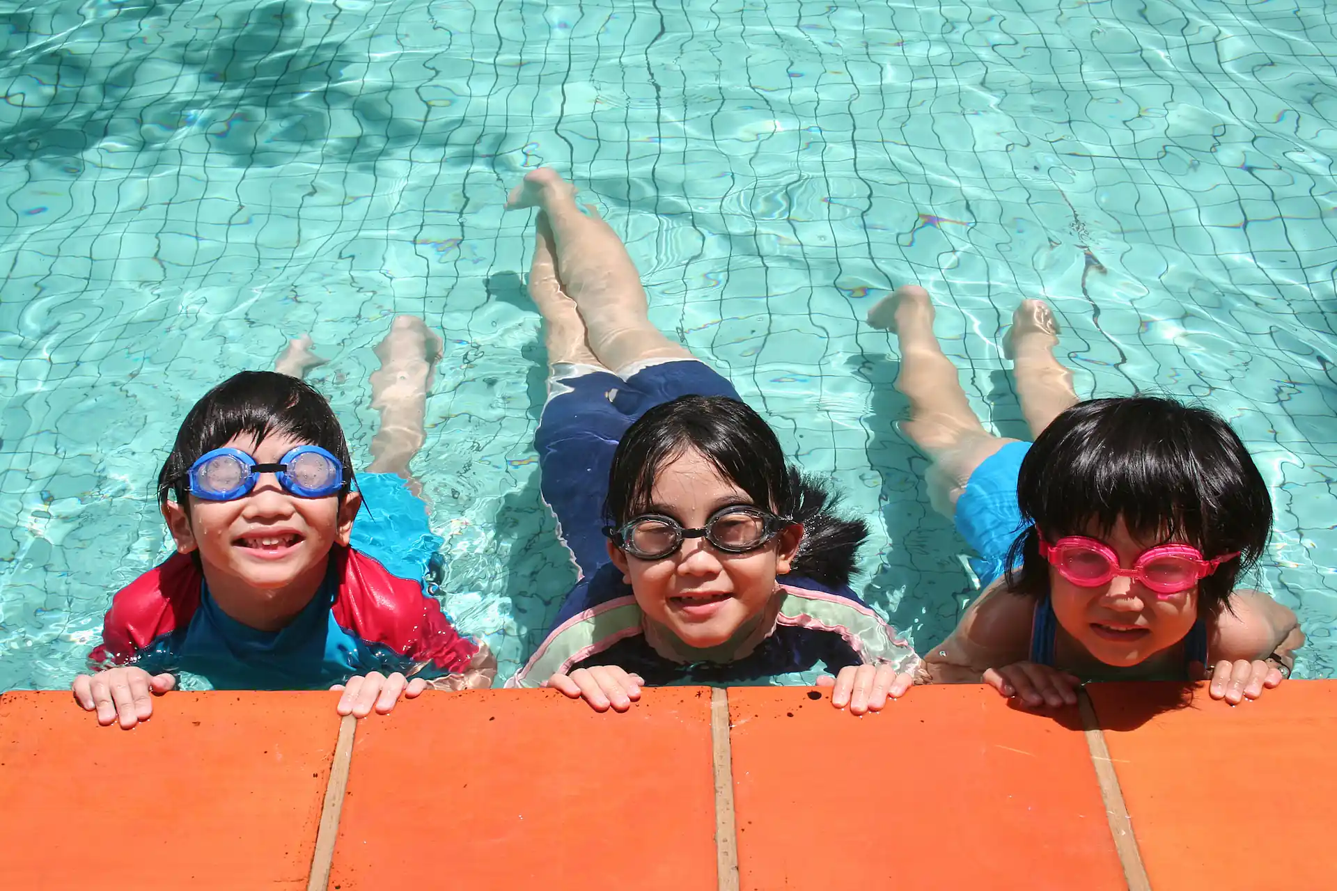 Three children in swimsuits and goggles smile while gripping the wall of a pool's shallow end. This vantage point helps demonstrate how to supervise your kids when they swim.