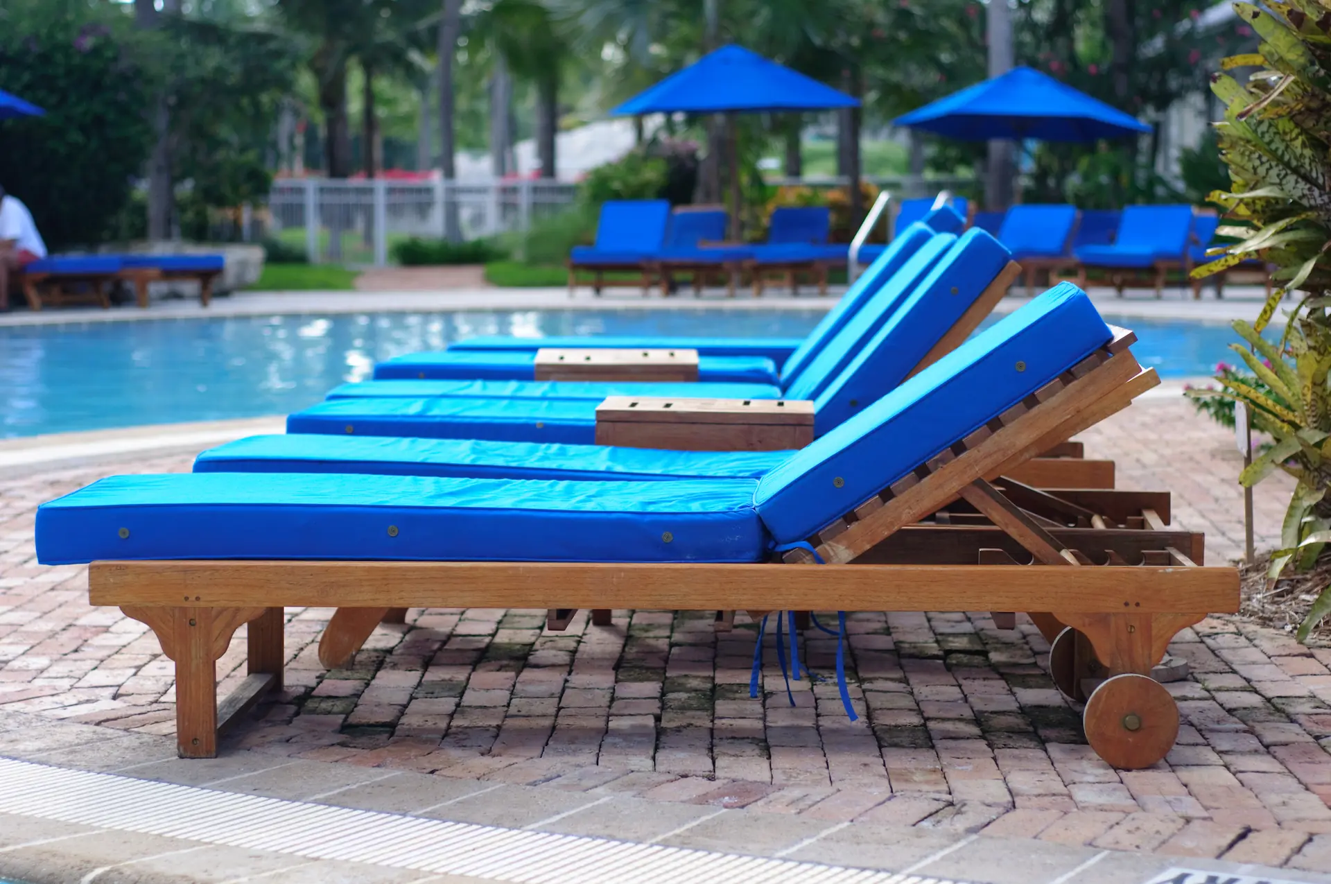 Profile of a row of wooden poolside lounge chairs with blue cushions. These are one of the options to consider in the in-pool chairs vs. poolside lounge chairs decision.